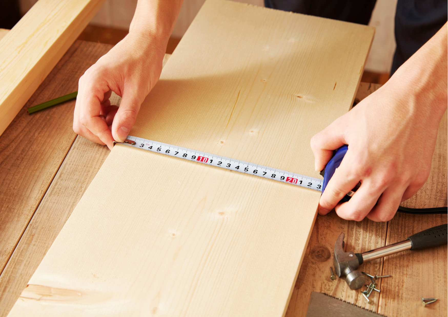 Mains d'un menuisier mesurant une planche de bois clair avec un mètre ruban sur un établi.
