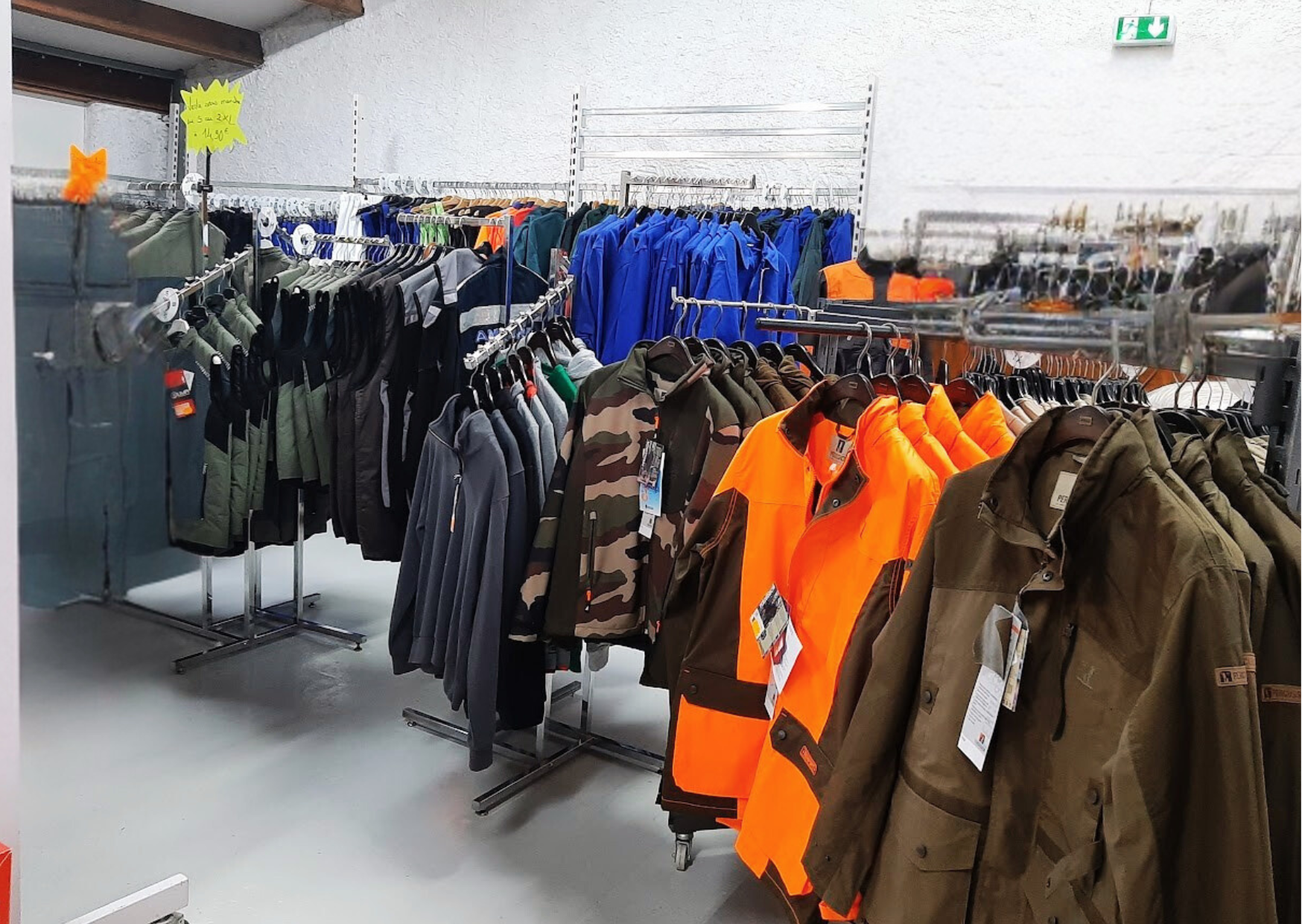 Rayonnage en magasin présentant des vestes de chasse camouflage, des équipements orange haute visibilité et des vêtements de travail professionnels.
