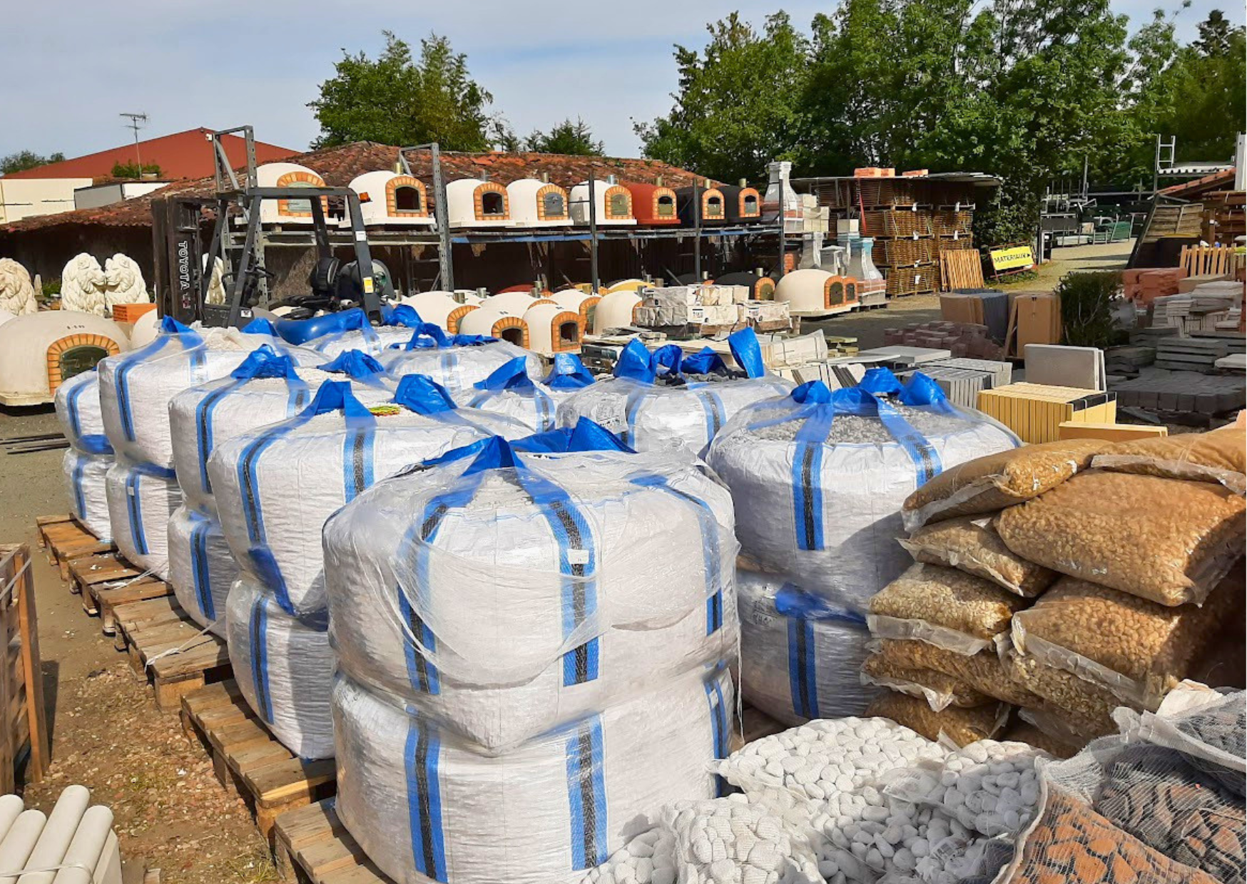 Big bags de matériaux en vrac, sable, graviers et alignement de fours à pizza traditionnels en stock chez Marais Fleuri.